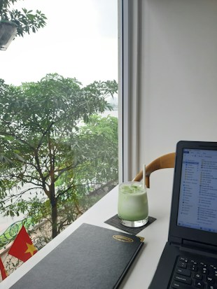 A modern workspace setup by a bright window featuring a lush green view outside with leafy trees. On the desk, there's a glass of iced green drink with a straw, a closed black notebook, and an open laptop displaying file windows. The Vietnamese flag is visible outside, adding a touch of national identity.