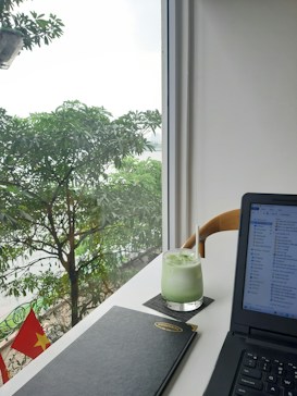 A modern workspace setup by a bright window featuring a lush green view outside with leafy trees. On the desk, there's a glass of iced green drink with a straw, a closed black notebook, and an open laptop displaying file windows. The Vietnamese flag is visible outside, adding a touch of national identity.