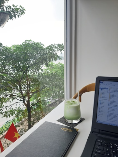 A modern workspace setup by a bright window featuring a lush green view outside with leafy trees. On the desk, there's a glass of iced green drink with a straw, a closed black notebook, and an open laptop displaying file windows. The Vietnamese flag is visible outside, adding a touch of national identity.