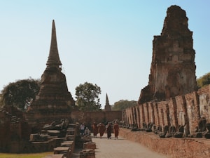 A group of Buddhist monks wearing traditional orange robes walks through ancient temple ruins. The scene features tall, pointed stupas and statues, with a clear blue sky above. The architecture is made of aged, reddish-brown bricks, and the area is surrounded by some trees.