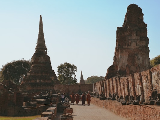 A group of Buddhist monks wearing traditional orange robes walks through ancient temple ruins. The scene features tall, pointed stupas and statues, with a clear blue sky above. The architecture is made of aged, reddish-brown bricks, and the area is surrounded by some trees.