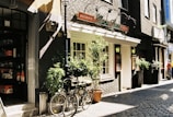 A cozy street with a brick building housing an Italian restaurant named 'Mama Leone'. There are two potted plants and a bicycle parked in front. To the left, part of a shop entrance with various items on display is visible. A person wearing a red shirt is walking further down the cobblestone path.