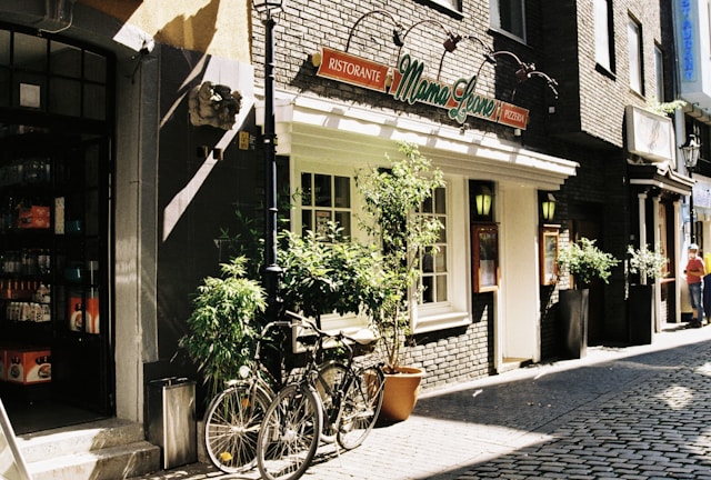 A cozy street with a brick building housing an Italian restaurant named 'Mama Leone'. There are two potted plants and a bicycle parked in front. To the left, part of a shop entrance with various items on display is visible. A person wearing a red shirt is walking further down the cobblestone path.