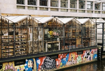 A row of greenhouses along a water canal with their glass walls displaying various dried flowers, fresh flowers, and bulbs. Below the greenhouses, a vibrant graffiti mural covers the wall, featuring stylized characters and vivid colors. The building in the background features rectangular windows in a grid pattern.