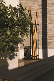 A decorative arrangement of tall bamboo sticks is placed in a black metal holder near a textured stone wall. Beside it, there is a dark glass vase with some plant stems. A leafy green plant partially obscures the view, and sunlight casts shadows on the wall and floor.