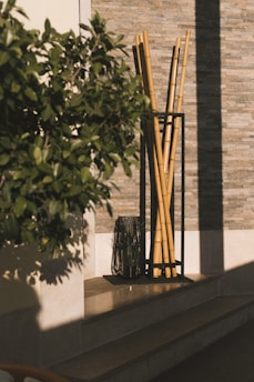 A decorative arrangement of tall bamboo sticks is placed in a black metal holder near a textured stone wall. Beside it, there is a dark glass vase with some plant stems. A leafy green plant partially obscures the view, and sunlight casts shadows on the wall and floor.
