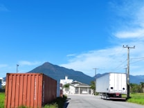 A scenic view featuring a large shipping container on the left, a semi-trailer truck on the right, and a mountain range under a clear blue sky. The road leads towards a house and green fields are visible along the sides.