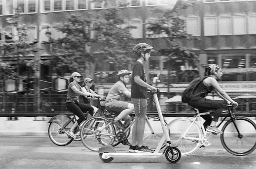 A black and white photograph featuring individuals on bicycles and a scooter in motion, likely on a city street. The people appear to be wearing helmets and casual clothing, moving in a line from left to right. Background buildings with windows and a hint of trees suggest an urban environment.
