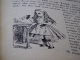 An illustration of a young girl holding a bottle labeled 'Drink Me', standing in front of a table. The image is part of a page from a book with visible text around the drawing.