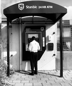A person is using an ATM outside, positioned in front of a Stanbic 24-hour cash dispenser. The individual is dressed in a white shirt and dark pants, and the setting is an outdoor area with a paved ground and nearby plants. The ATM is shaded by a canopy bearing the bank's logo and name.