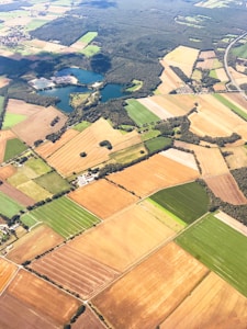 Aerial view of a rural landscape featuring a patchwork of rectangular agricultural fields in various shades of green, brown, and yellow. A small lake is surrounded by dense forest areas. Roads and small clusters of buildings dot the countryside.