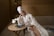 A woman in a white bathrobe and towel wrapped around her head sits on a sofa in a spa-like setting. She is reading a document, with a table in front of her holding a vase with white flowers, a candle, and a magazine. The room has wooden walls, creating a warm, relaxing environment.