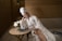 A woman in a white bathrobe and towel wrapped around her head sits on a sofa in a spa-like setting. She is reading a document, with a table in front of her holding a vase with white flowers, a candle, and a magazine. The room has wooden walls, creating a warm, relaxing environment.