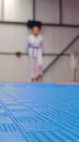 A blurry image with a focus on a blue mat in the foreground, while in the background, a figure wearing what appears to be a martial arts uniform is faintly visible standing in front of a wall with vertical and diagonal lines.
