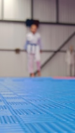 A blurry image with a focus on a blue mat in the foreground, while in the background, a figure wearing what appears to be a martial arts uniform is faintly visible standing in front of a wall with vertical and diagonal lines.
