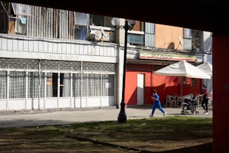 A city street scene featuring a beauty salon and a pizzeria with bright red walls. A lamppost stands in the foreground. People are visible; one person in blue is walking, while another person pushes a stroller, both wearing masks. The outdoor seating area of the pizzeria is shaded by a large umbrella.