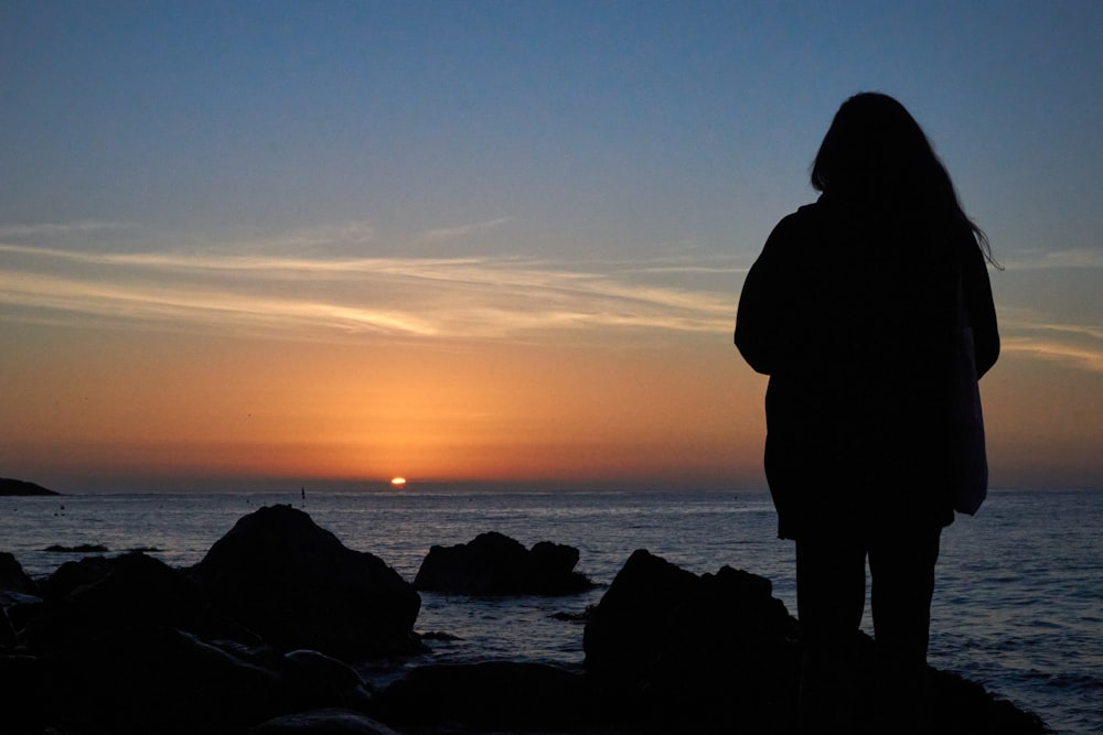 silhouette of person standing on rock formation near body of water during sunset