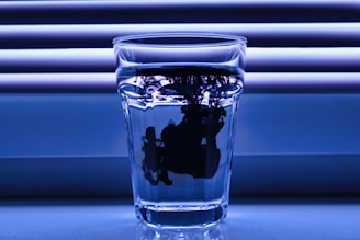 A clear glass filled with water stands against a background of horizontal blinds. A dark, ink-like substance is diffusing within the water, creating intricate and abstract shapes.