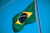 A Brazilian flag is waving against a clear blue sky. The flag includes a green field with a yellow diamond in the center, which encases a blue circle adorned with white stars and the motto 'Ordem e Progresso'.