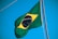 A Brazilian flag is waving against a clear blue sky. The flag includes a green field with a yellow diamond in the center, which encases a blue circle adorned with white stars and the motto 'Ordem e Progresso'.