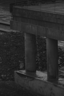A partially constructed concrete bridge with large support pillars, surrounded by gravel and dirt. The structure lacks finishing elements and the setting appears overcast and industrial.