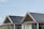 A building with large windows and sloped roofs covered in dark solar panels. A seagull is perched on the peak of one of the rooftops. The background features a clear sky and grassy area, with distant buildings partially visible.