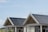 A building with large windows and sloped roofs covered in dark solar panels. A seagull is perched on the peak of one of the rooftops. The background features a clear sky and grassy area, with distant buildings partially visible.