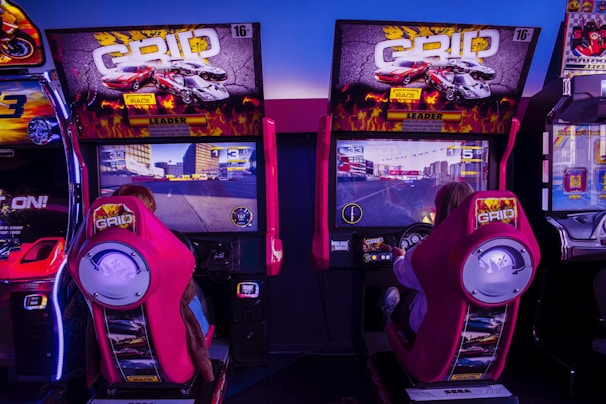 Two people are seated in an arcade racing setup, playing a car racing game called GRID. They are sitting in red racing seats with steering wheels in front of large screens displaying the racing game. The arcade machines are surrounded by bright neon lights and bold graphics, creating an immersive gaming environment.