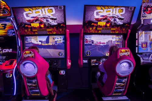 Two people are seated in an arcade racing setup, playing a car racing game called GRID. They are sitting in red racing seats with steering wheels in front of large screens displaying the racing game. The arcade machines are surrounded by bright neon lights and bold graphics, creating an immersive gaming environment.