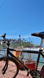 A bicycle is parked on a promenade with a red and white lighthouse in the background. The scene includes a clear blue sky and the ocean waves crashing against the rocky shore lined with palm trees. There are also potted plants along the path.