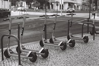 A row of electric scooters is parked on a tiled sidewalk next to a street. The scooters are neatly arranged and appear to be part of a rental service. The street shows typical urban elements like cars, pedestrians, and buildings in the background.