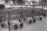 A row of electric scooters is parked on a tiled sidewalk next to a street. The scooters are neatly arranged and appear to be part of a rental service. The street shows typical urban elements like cars, pedestrians, and buildings in the background.