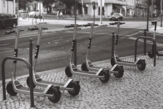 A row of electric scooters is parked on a tiled sidewalk next to a street. The scooters are neatly arranged and appear to be part of a rental service. The street shows typical urban elements like cars, pedestrians, and buildings in the background.