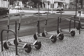 A row of electric scooters is parked on a tiled sidewalk next to a street. The scooters are neatly arranged and appear to be part of a rental service. The street shows typical urban elements like cars, pedestrians, and buildings in the background.