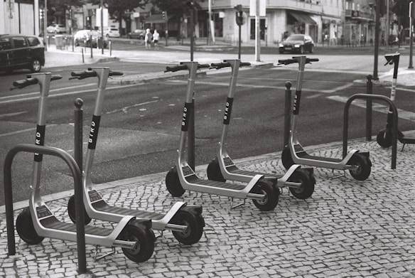 A row of electric scooters is parked on a tiled sidewalk next to a street. The scooters are neatly arranged and appear to be part of a rental service. The street shows typical urban elements like cars, pedestrians, and buildings in the background.