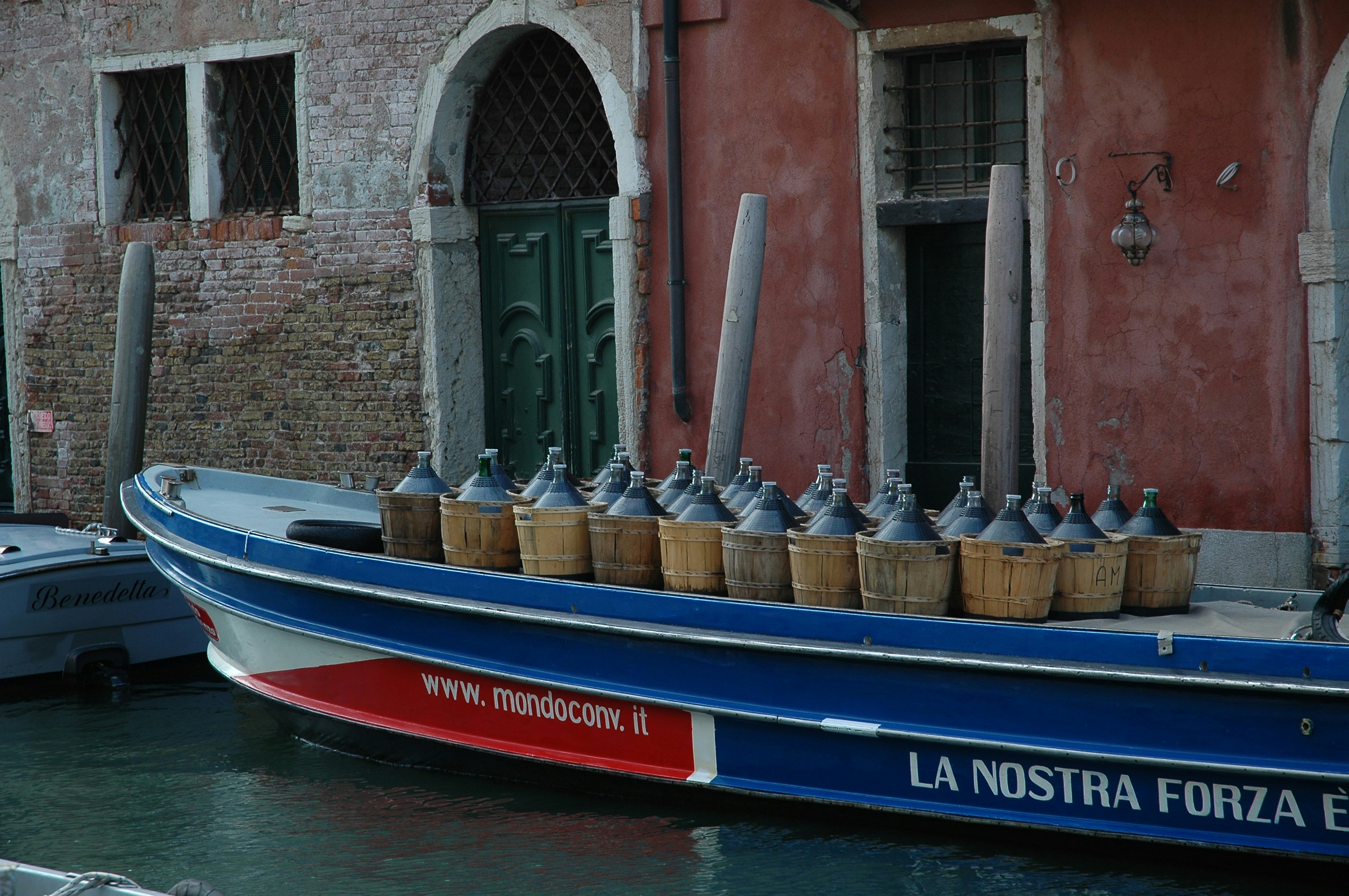 A traditional Venetian canal boat is docked alongside an aged brick building with a rustic red facade. The boat is loaded with large glass demijohns, each encased in wicker or wooden carriers. The building features arched doorways and small barred windows, adding to the historic charm.