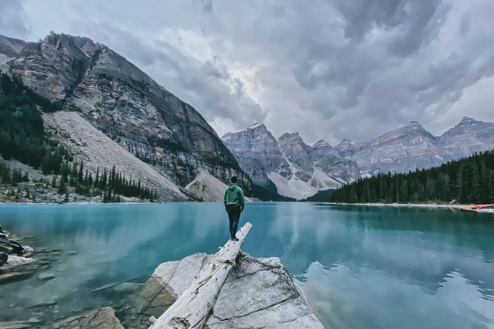 person in green jacket and black pants standing on rock near lake during daytime