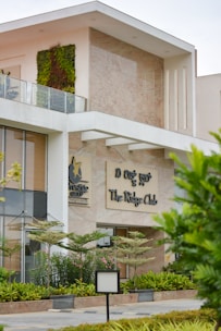 A modern building with a partially visible green wall on the upper level and large glass windows. The sign reads 'The Ridge Club' and is accompanied by the logo of the Prestige Group. The facade features contemporary architecture with clean lines and a mix of stone and glass. There are landscaped plants and trees in the front area, adding a touch of greenery.