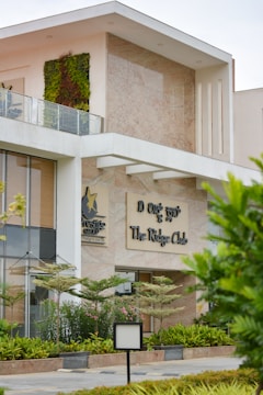 A modern building with a partially visible green wall on the upper level and large glass windows. The sign reads 'The Ridge Club' and is accompanied by the logo of the Prestige Group. The facade features contemporary architecture with clean lines and a mix of stone and glass. There are landscaped plants and trees in the front area, adding a touch of greenery.