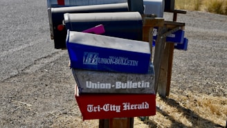 Several mailboxes are stationed along a gravel road, with most featuring newspaper names and logos. The mailboxes have a worn, rustic appearance and are mounted on a wooden frame. The ground is covered with dry grass.