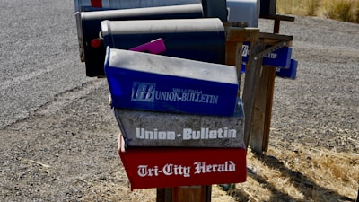 Several mailboxes are stationed along a gravel road, with most featuring newspaper names and logos. The mailboxes have a worn, rustic appearance and are mounted on a wooden frame. The ground is covered with dry grass.
