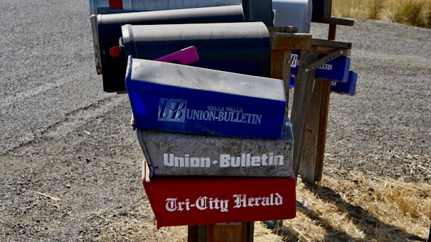 Several mailboxes are stationed along a gravel road, with most featuring newspaper names and logos. The mailboxes have a worn, rustic appearance and are mounted on a wooden frame. The ground is covered with dry grass.