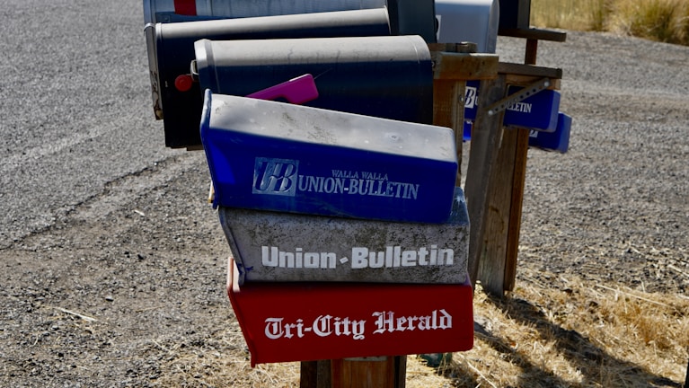 Several mailboxes are stationed along a gravel road, with most featuring newspaper names and logos. The mailboxes have a worn, rustic appearance and are mounted on a wooden frame. The ground is covered with dry grass.