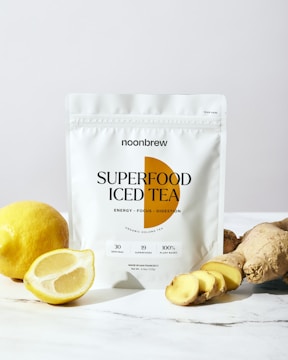A white package labeled 'Superfood Iced Tea' is prominently displayed on a marble surface. To the left of the package, a whole lemon and a halved lemon are visible. To the right, several pieces of ginger root are arranged, some of which are sliced. The composition suggests freshness and healthiness, with light highlighting the natural ingredients.