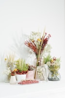 Multiple arrangements of dried flowers and plants are displayed in various vases and containers. The vases are transparent and ceramic, holding different varieties of greenery and colorful dried flowers, including reds, yellows, and whites. The setup is placed on a white table against a light-colored wall, giving it an elegant and natural look.