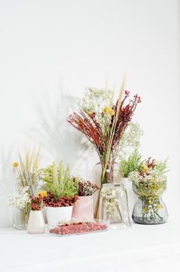 Multiple arrangements of dried flowers and plants are displayed in various vases and containers. The vases are transparent and ceramic, holding different varieties of greenery and colorful dried flowers, including reds, yellows, and whites. The setup is placed on a white table against a light-colored wall, giving it an elegant and natural look.