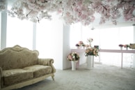 A room with an elegant floral theme, featuring a vintage-style couch with intricate patterns. Numerous flower arrangements in vases are placed on a table and on the floor. Overhead, a canopy of white and pink artificial blossoms adds a dreamy essence. Natural light filters in through large windows, creating a serene and airy atmosphere.
