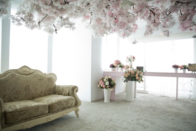 A room with an elegant floral theme, featuring a vintage-style couch with intricate patterns. Numerous flower arrangements in vases are placed on a table and on the floor. Overhead, a canopy of white and pink artificial blossoms adds a dreamy essence. Natural light filters in through large windows, creating a serene and airy atmosphere.