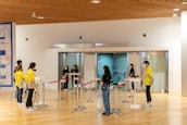A group of people are gathered in a large hallway with a wooden ceiling. Some individuals are wearing yellow shirts labeled 'staff' and 'volunteer,' while others are dressed in casual clothing. Several are wearing face masks, suggesting a public setting. There are red ropes set up for line management and a digital check-in point at the entrance to a room. A large mural or poster can be seen in the background.