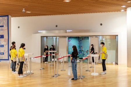 A group of people are gathered in a large hallway with a wooden ceiling. Some individuals are wearing yellow shirts labeled 'staff' and 'volunteer,' while others are dressed in casual clothing. Several are wearing face masks, suggesting a public setting. There are red ropes set up for line management and a digital check-in point at the entrance to a room. A large mural or poster can be seen in the background.